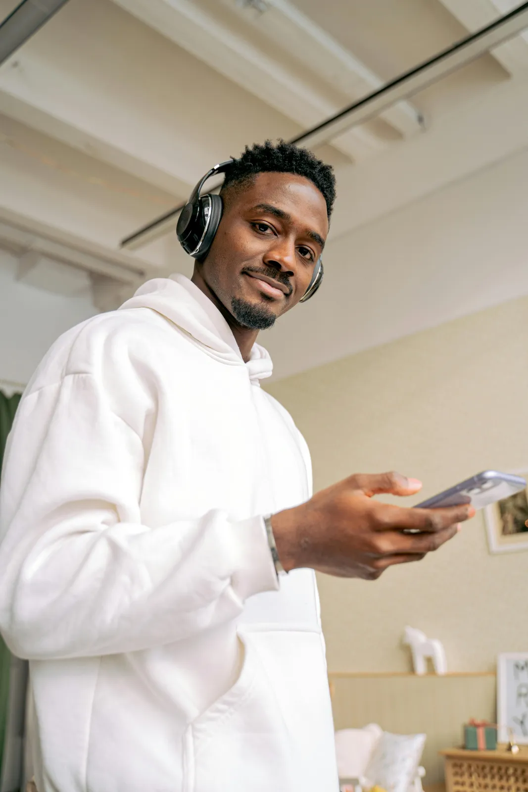 Ein Mann mit Kopfhörern und weißem Hoodie hält lächelnd sein Smartphone in der Hand.