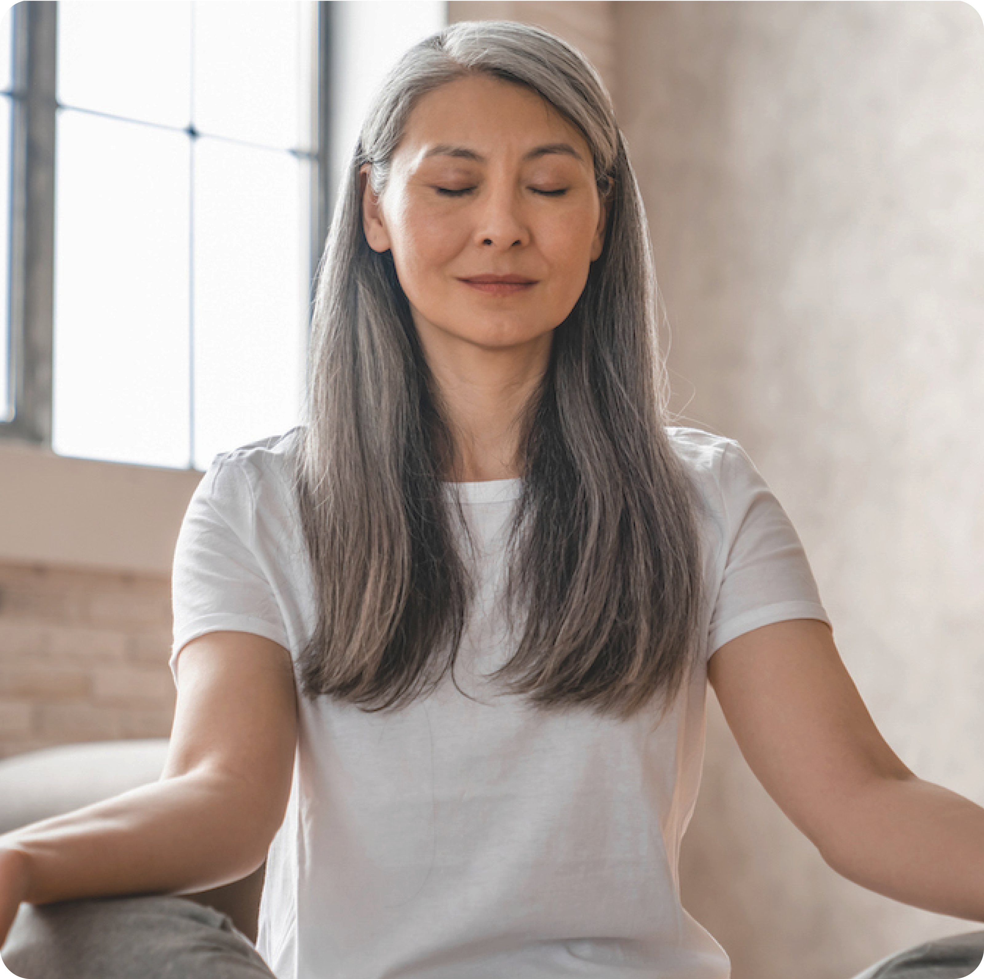 A woman sits upright in a bright room with her eyes closed