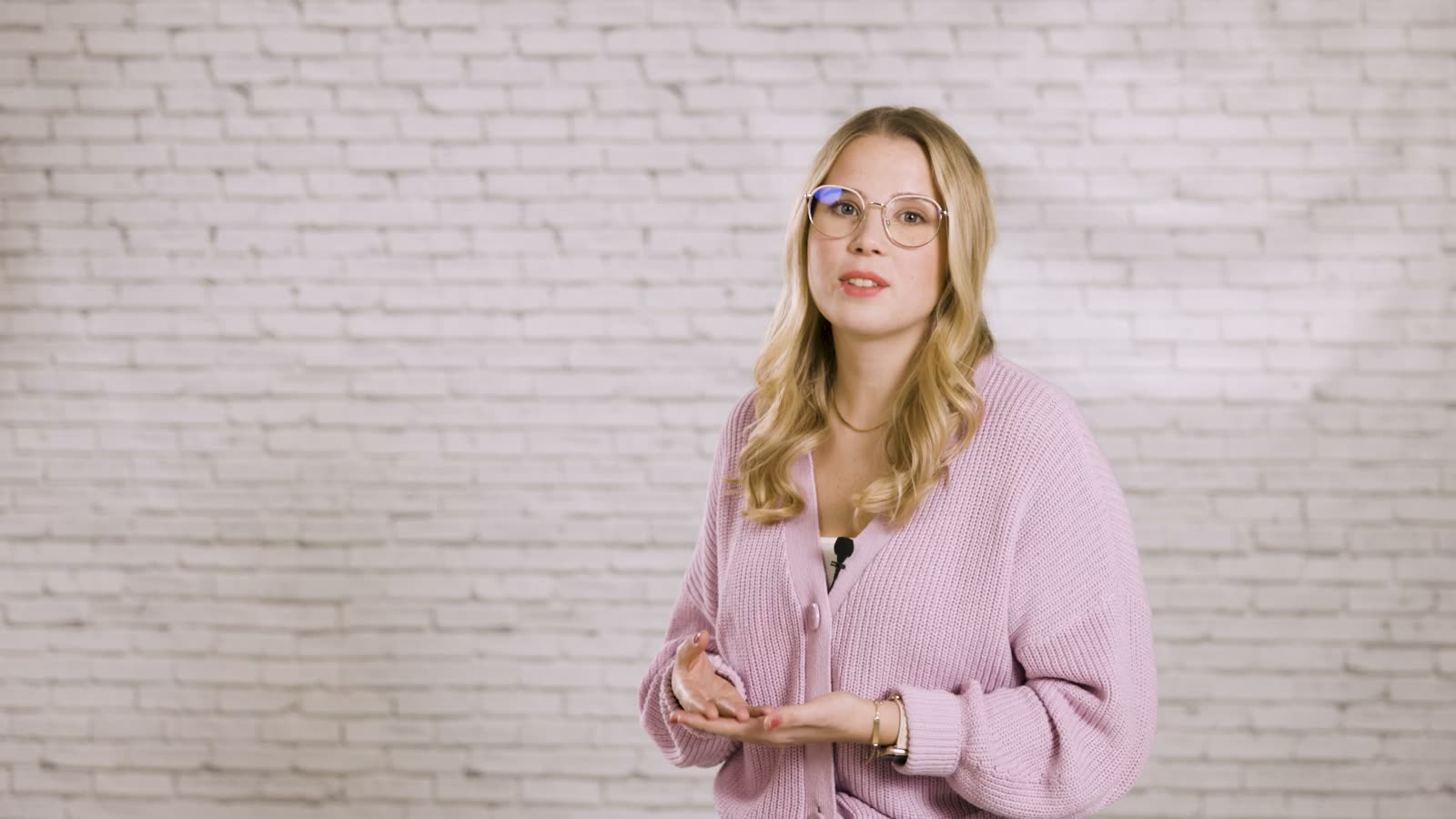 Eine junge Frau mit Brille und rosa Strickjacke spricht vor einer weißen Backsteinwand in die Kamera