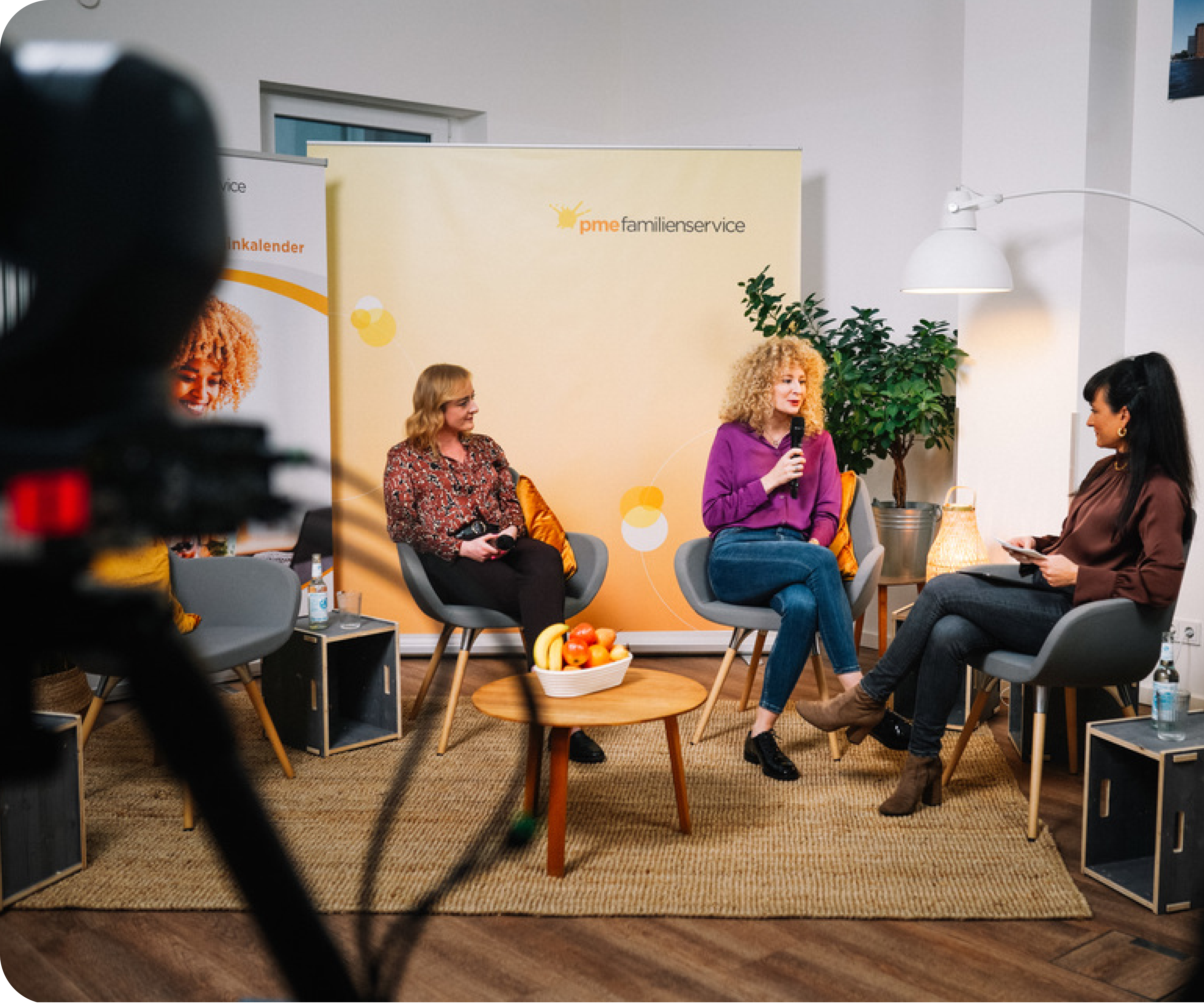 Drei Frauen sitzen in einem Studio im Gespräch, im Hintergrund ein Rollup des pme Familienservice, im Vordergrund eine Kamera
