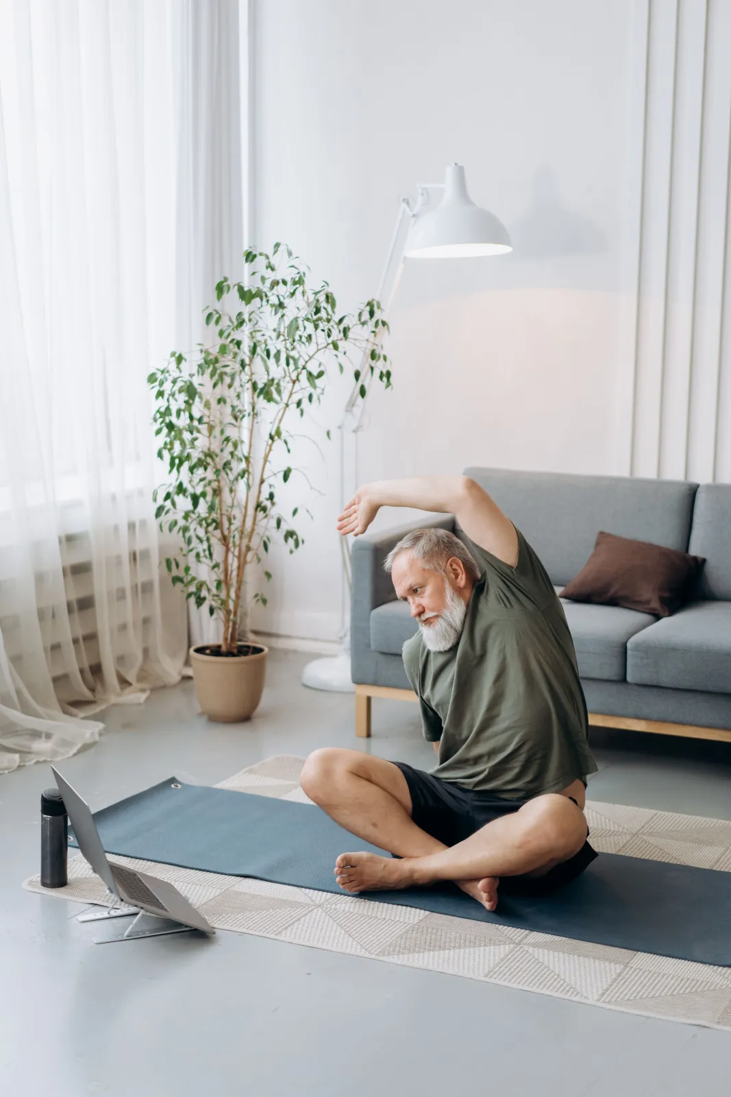 A man with a grey beard stretches on a yoga mat in his living room, following a live class on his laptop